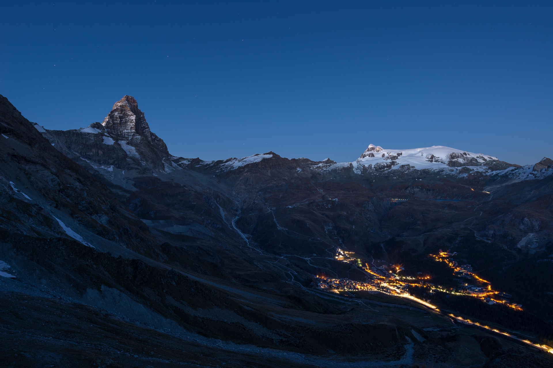Aerial view of Breuil Cervinia village glowing in the night, famous ski resort in Aosta Valley, Italy. Wonderful starry sky over Matterhorn (Cervino) mountain peak and Monte Rosa glaciers.